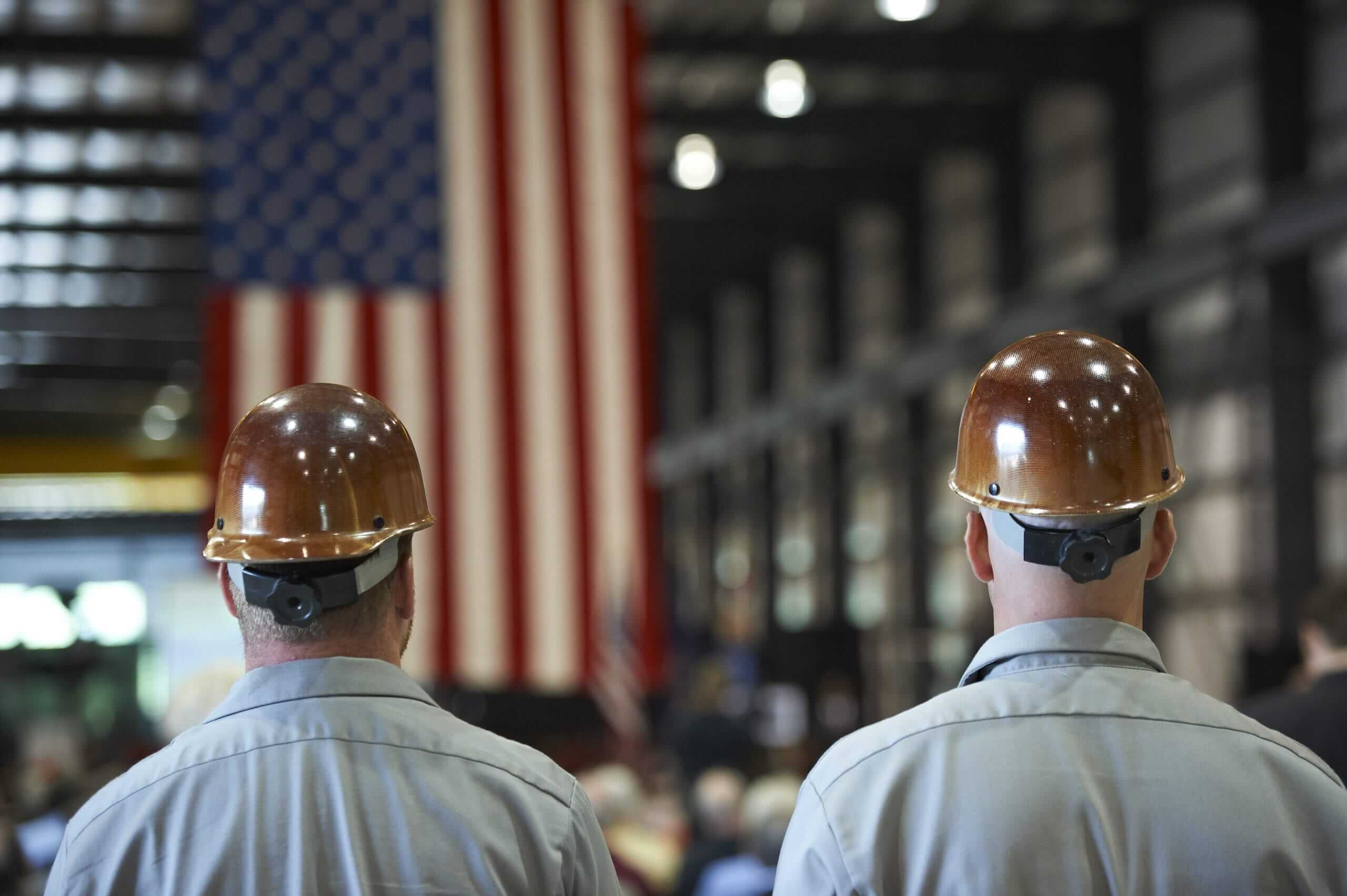 A photo of two men from the back wearing hard hats while looking at the American flag.