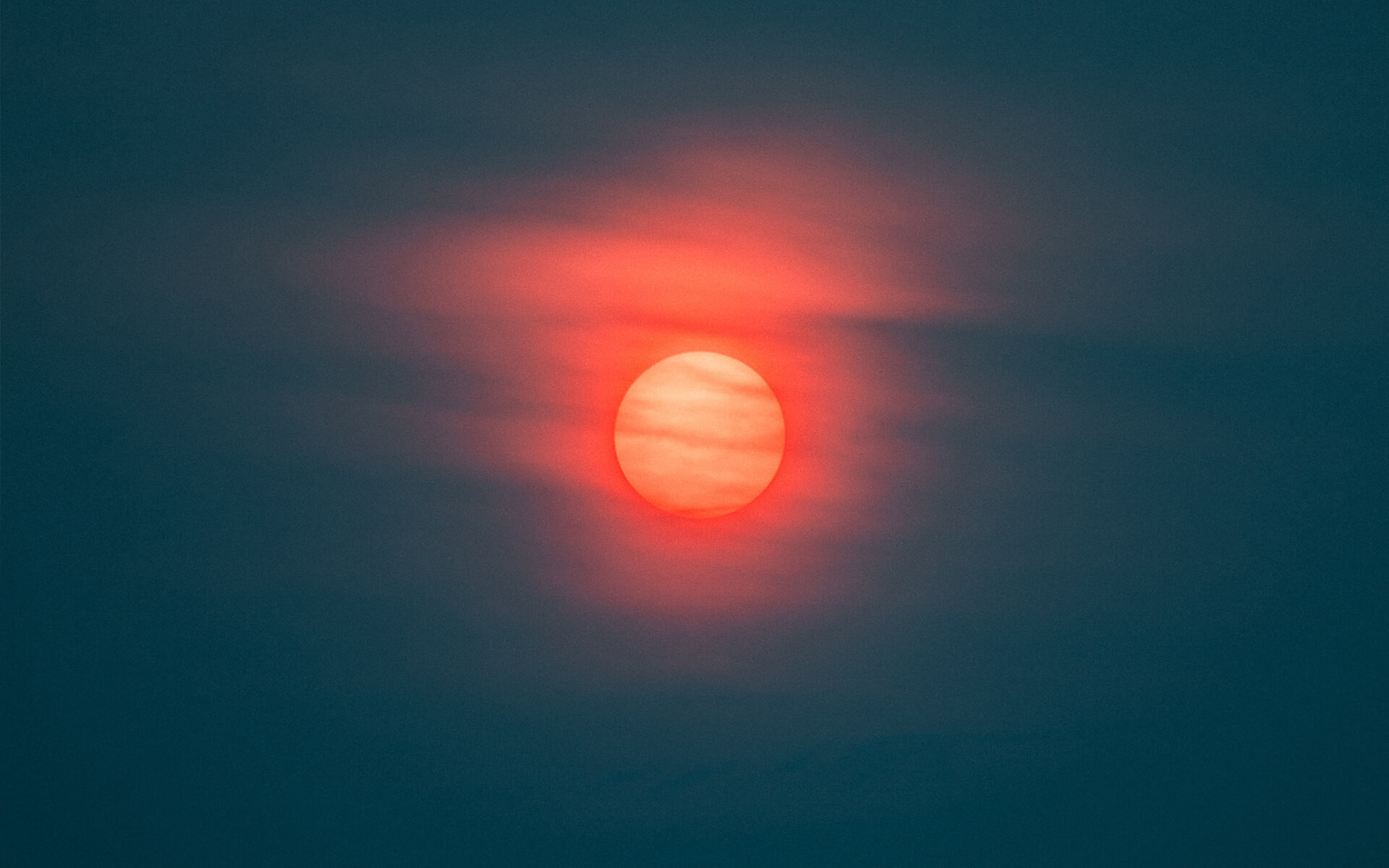 A photo of the sun producing red light against a dark blue sky, with clouds passing across its face.